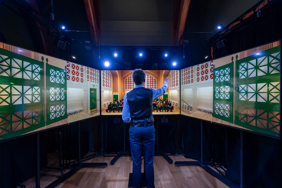 A male student practising talking in front of a smaller visual simulator, with a screen in front and two screens to the side.